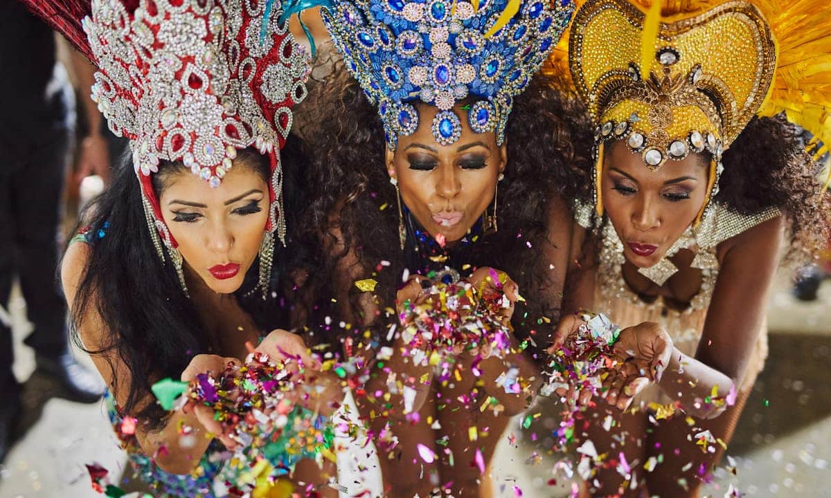 Three individuals in elaborate, colorful costumes with ornate headdresses adorned with feathers and jewels, blowing colorful confetti into the air during a festive celebration.