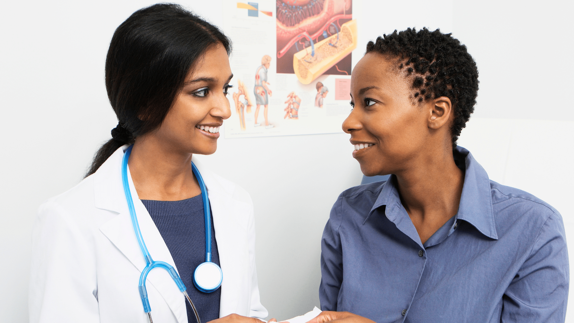 A medical professional in a white coat with a stethoscope around their neck engages in a conversation with another person in a clinical setting. A colorful anatomical chart on the wall in the background displays various medical diagrams, reflecting a focus on medical education and residency preparation at All Saints University.