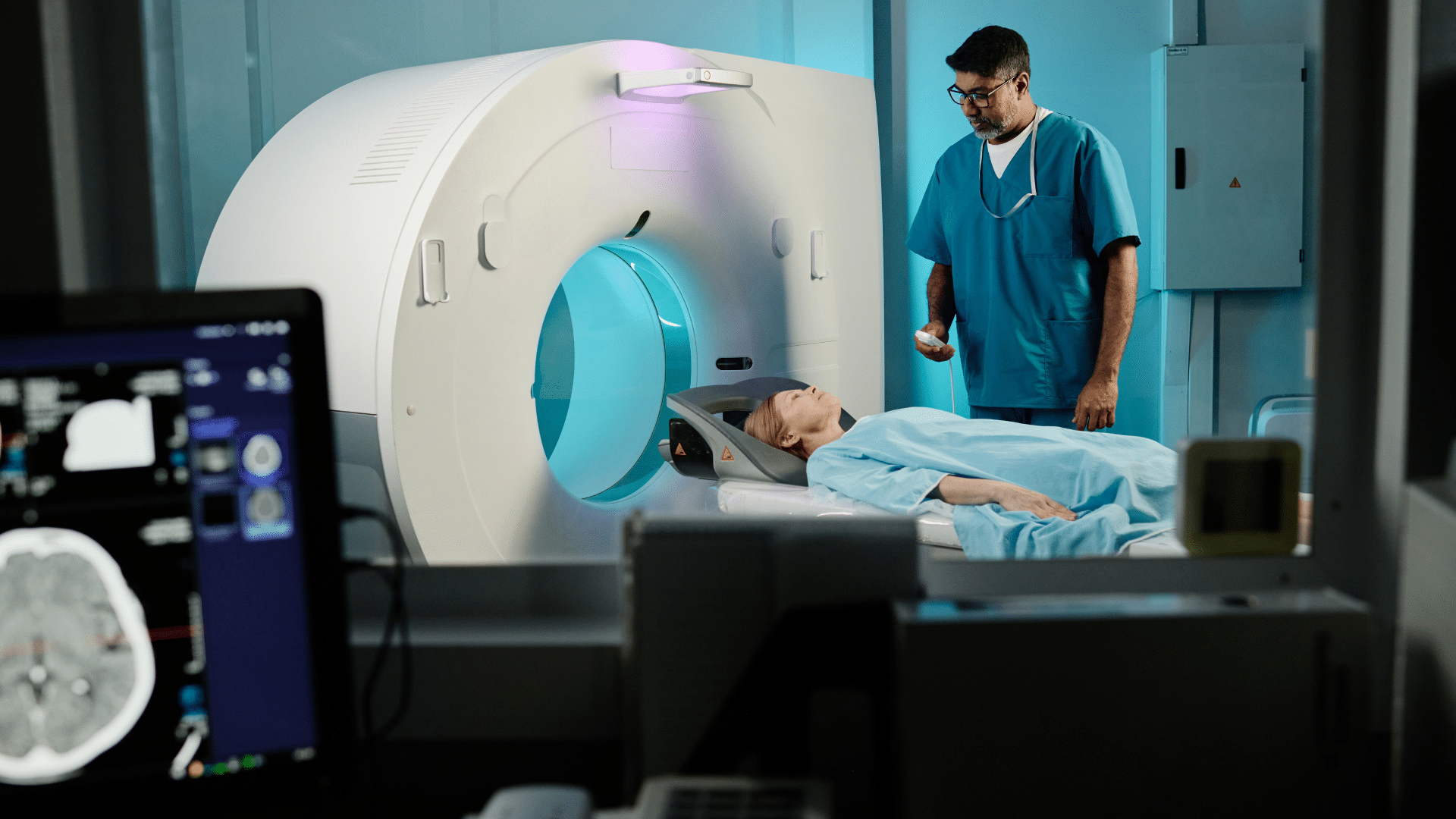 A patient lies on a table, partially inside a CT scanner, while a medical professional in blue scrubs stands nearby, holding a remote control device. A computer monitor in the foreground displays a brain scan image. The room has a clinical, blue-toned ambiance with medical equipment visible in the background.