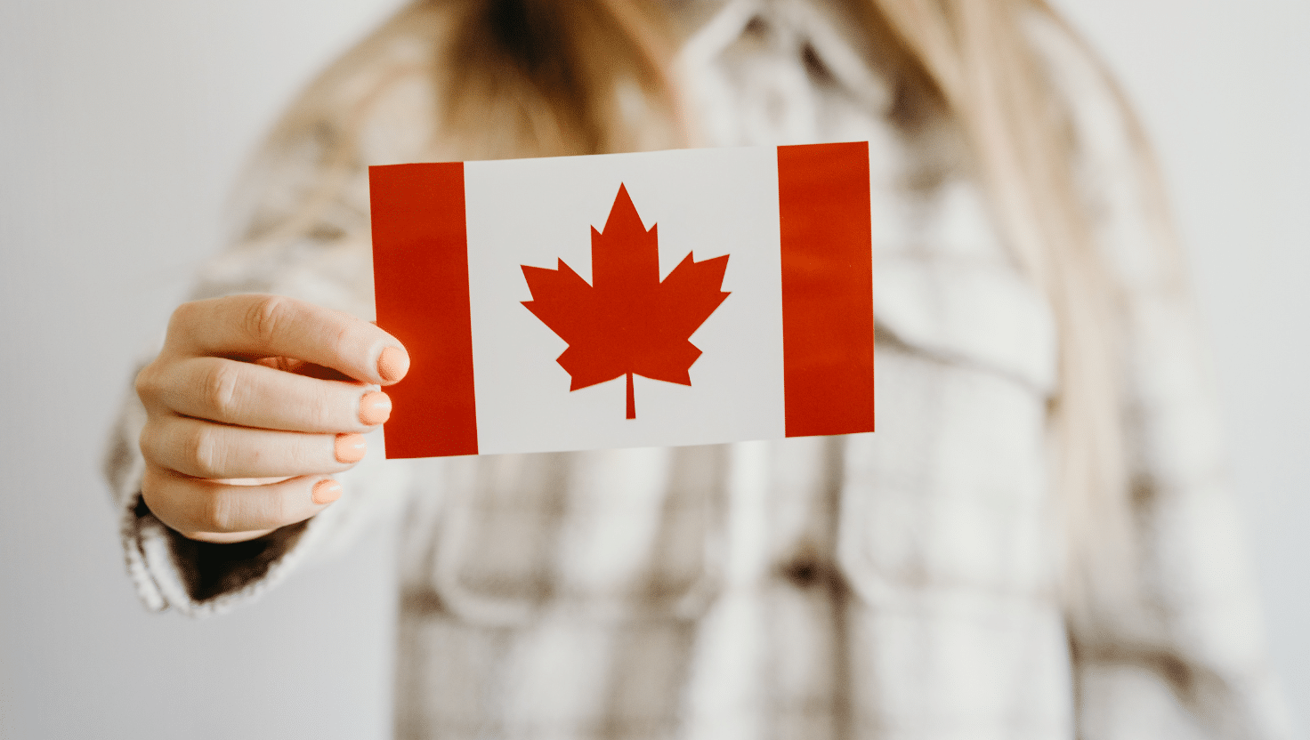 A person with long blonde hair holds a Canadian flag, featuring a red maple leaf and red vertical stripes on a white background, symbolizing opportunities for Canadian students at All Saints University.